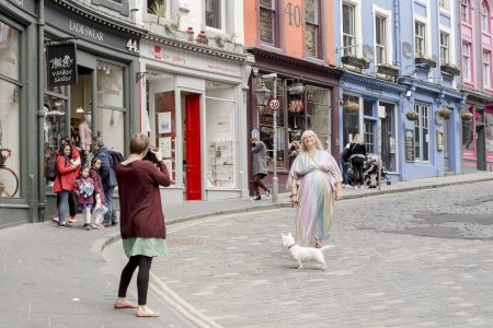 image shows a lady in a rainbow dress with pink boots and a small white dog, she is walking down a street in Edinburgh posing for her business headshots. In the foreground you can also see the photographer taking the photo.