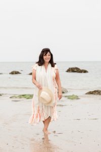 lady walking on the beach on a grey day wearing a sun dress and carrying her sun hat