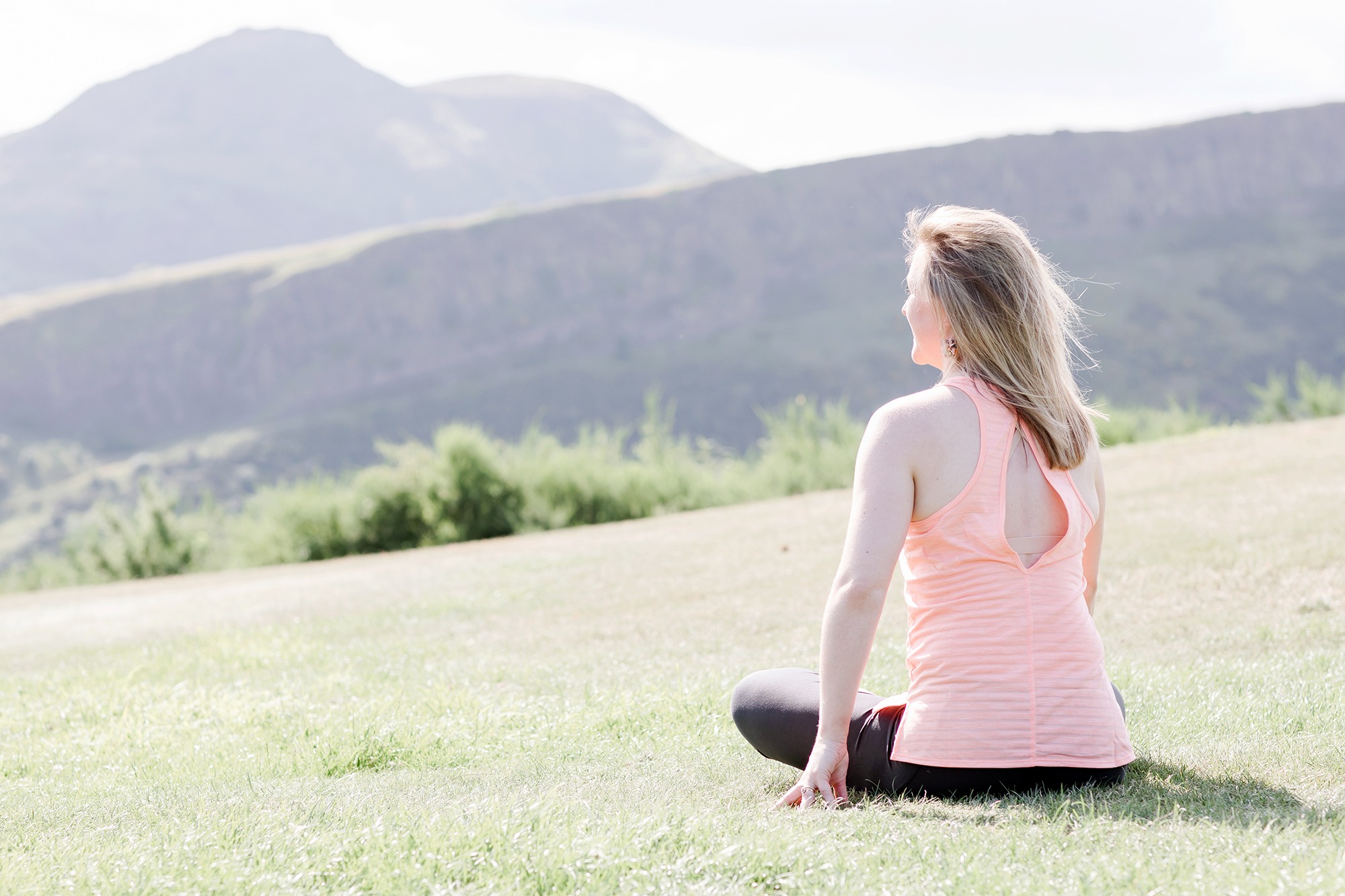 woman sitting cross legged on the grass with her back to the camera, looking out to hills